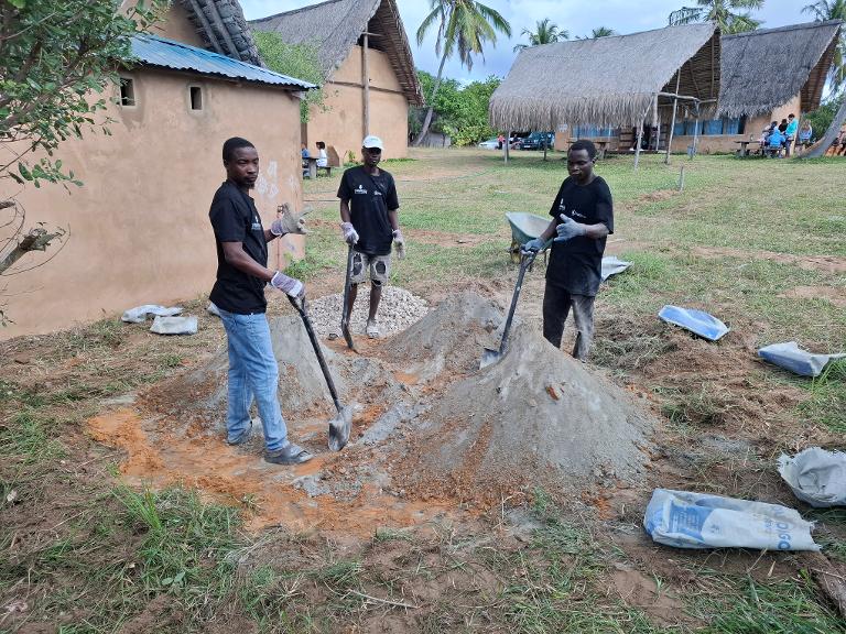 Josef, Augusto og Bonjon håndblander betong til forskaling. Kledd opp i t-skjorter fra Stavnesli byggservice.